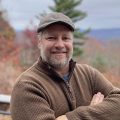 Man smiles with his hands crossed in a brown sweater. Trees and mountains in the fall are seen in the background.