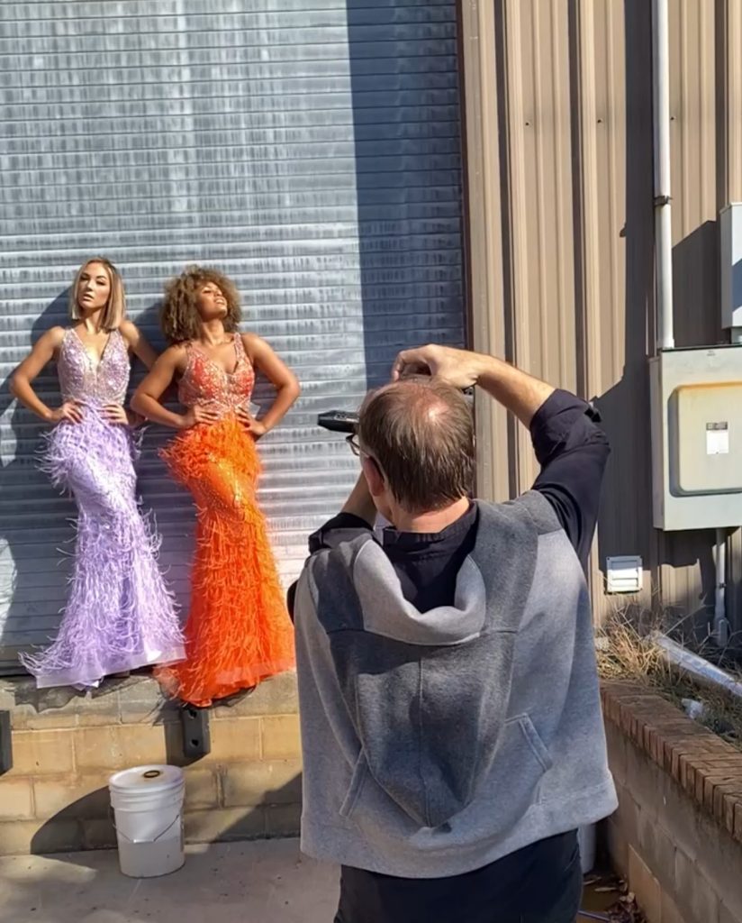 A man holding a camera (and visible from the back) takes a photo of two women wearing ball gowns who are leaning against a wall.
