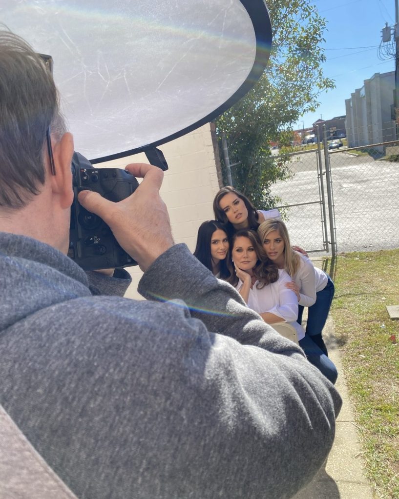 The corner of a man's arm is visible as he holds a camera and points it towards a group of four women wearing white shirts and posing for a photo outdoors.