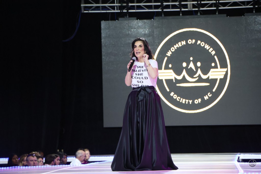 A white woman stands on a runway and speaks into a mic. She is wearing a T-shirt that has the words "She believed she could so she did" printed on it and a long black skirt.