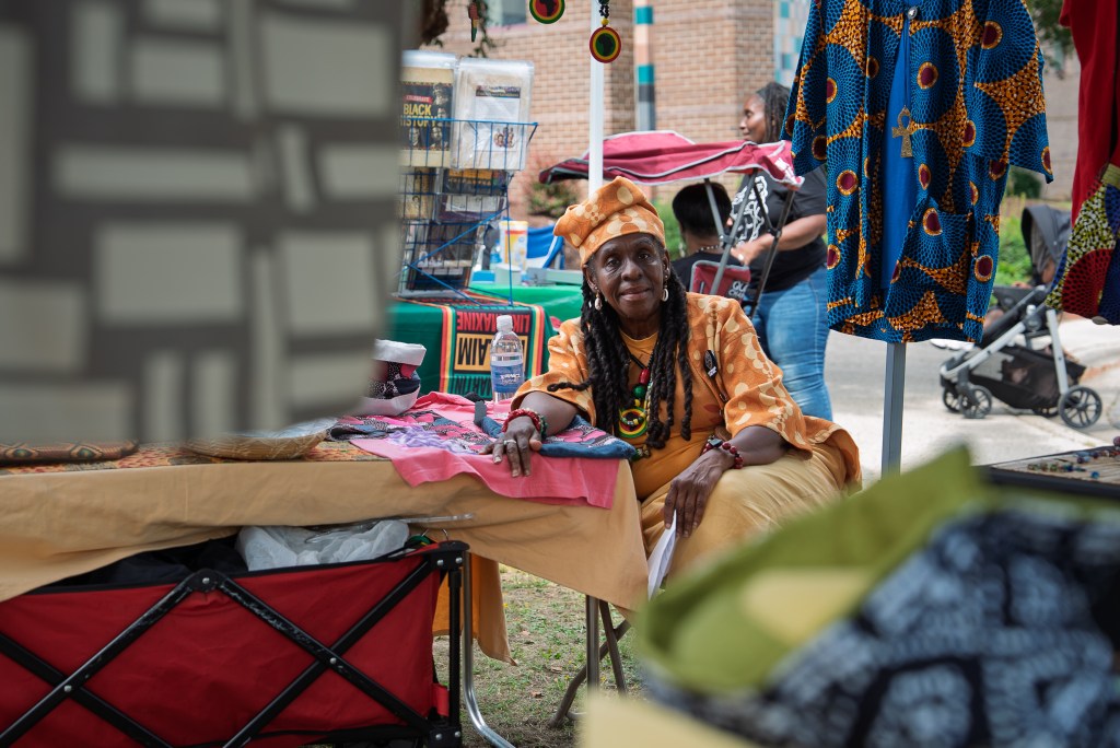 A Black woman in bright orange clothing leans against her vendor table at a festival and smiles at the camera.