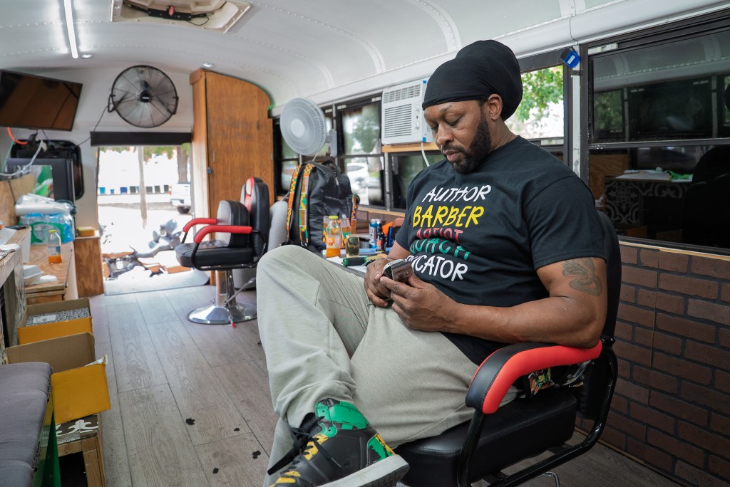A Black man stits in a barber chair inside a renovated school bus that serves as a mobile barber shop.
