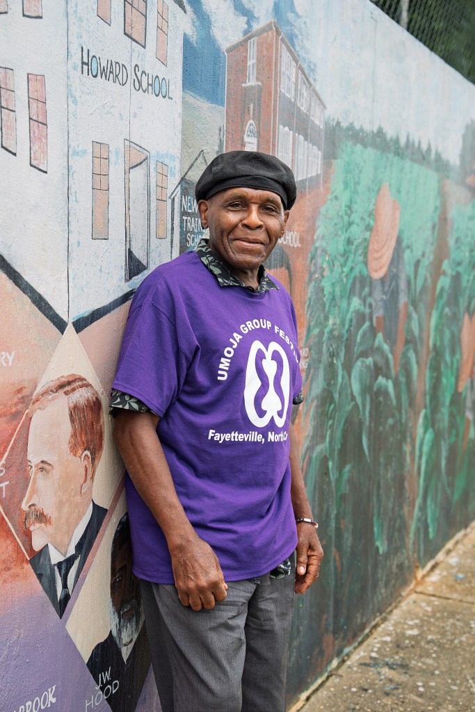 A Black man smiles at the camera for a portrait, wearing a bright purple shirt reading "Umoja Group Festival"