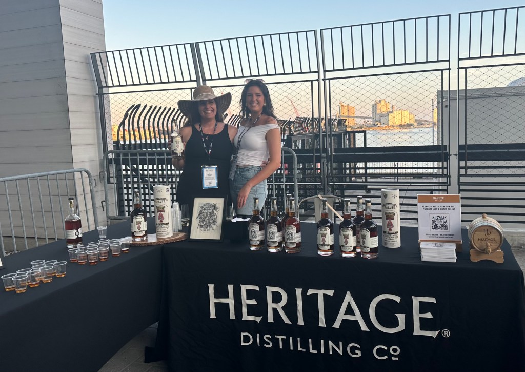 Two women smile and stand behind a table with a cloth draped over it reading "Heritage Distilling Co."